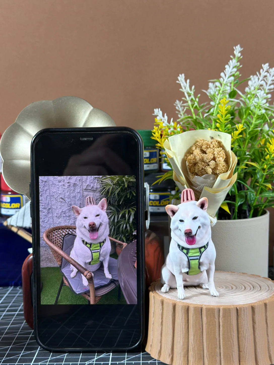 White dog reference photo on a phone beside a finished figurine with a green harness